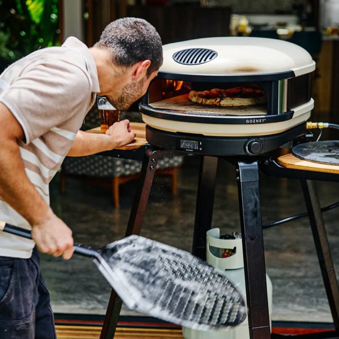 A man uses a pizza peel to handle food in a round, freestanding outdoor grill or pizza oven with a cream and black design, set on a dark metal stand with a visible propane tank underneath.